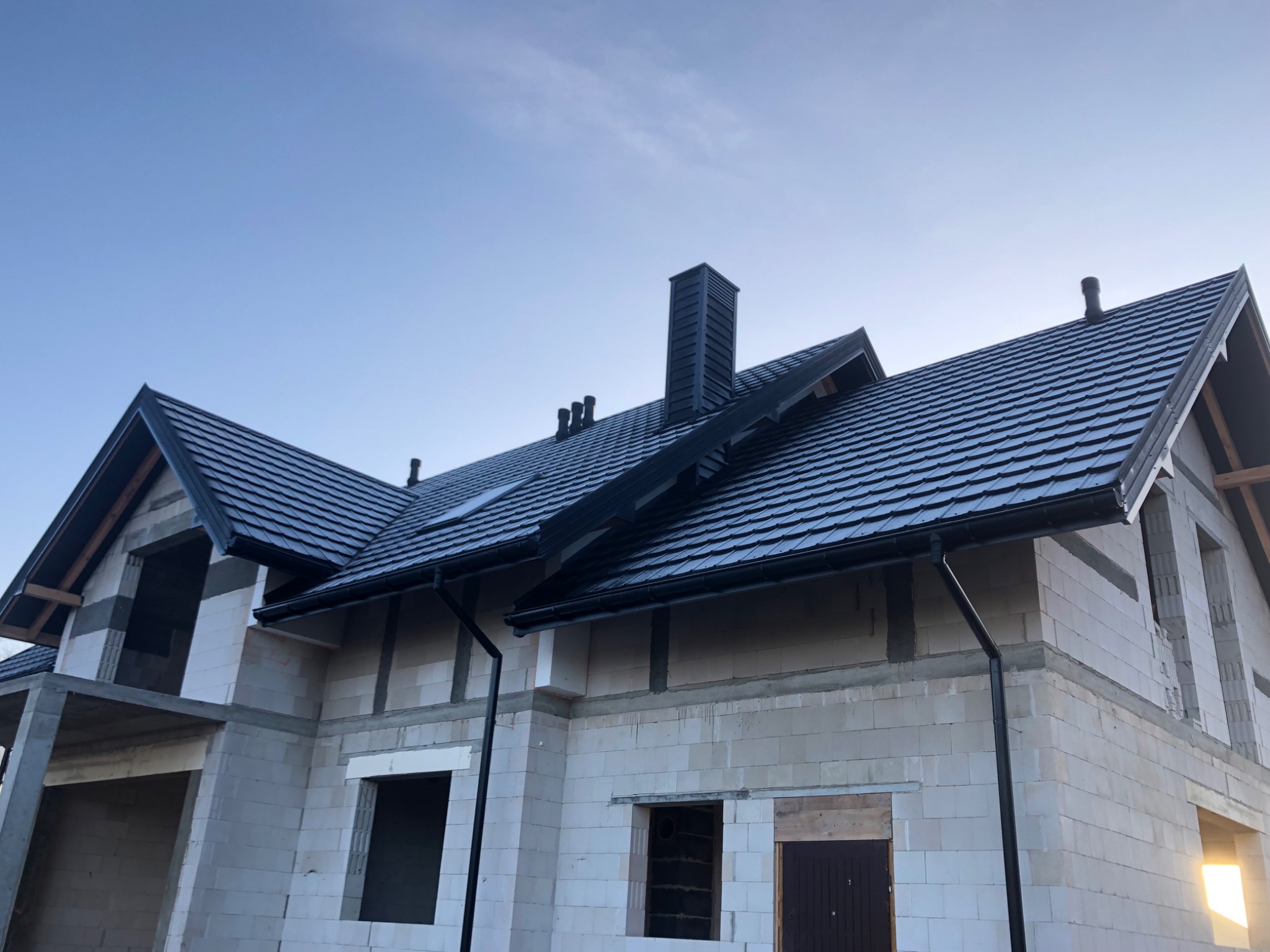 Niedokończony dom z białego pustaka z zamontowanym, czarnym dachem z dachówki ceramicznej, czarnymi rynnami i kominem, widoczne niezabezpieczone otwory okienne i drzwiowe.