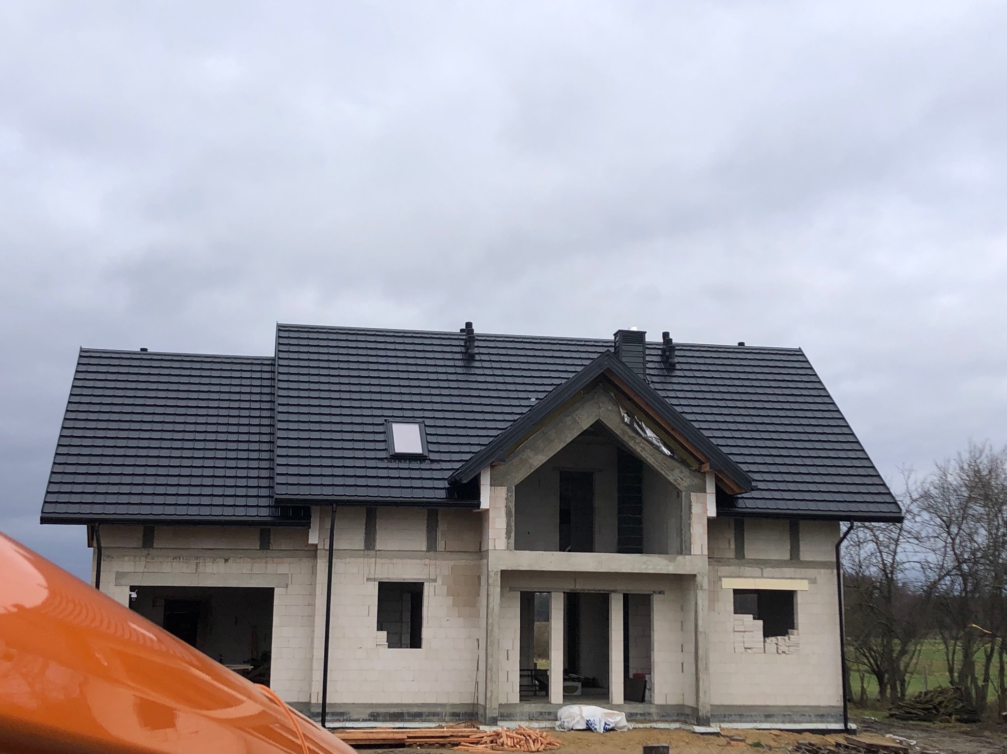 Dom w trakcie budowy z nowym, ciemnym dachem z dachówki ceramicznej i lukarną, widoczne niedokończone ściany z białego bloczka i pomarańczowy element dźwigu w lewym dolnym rogu.