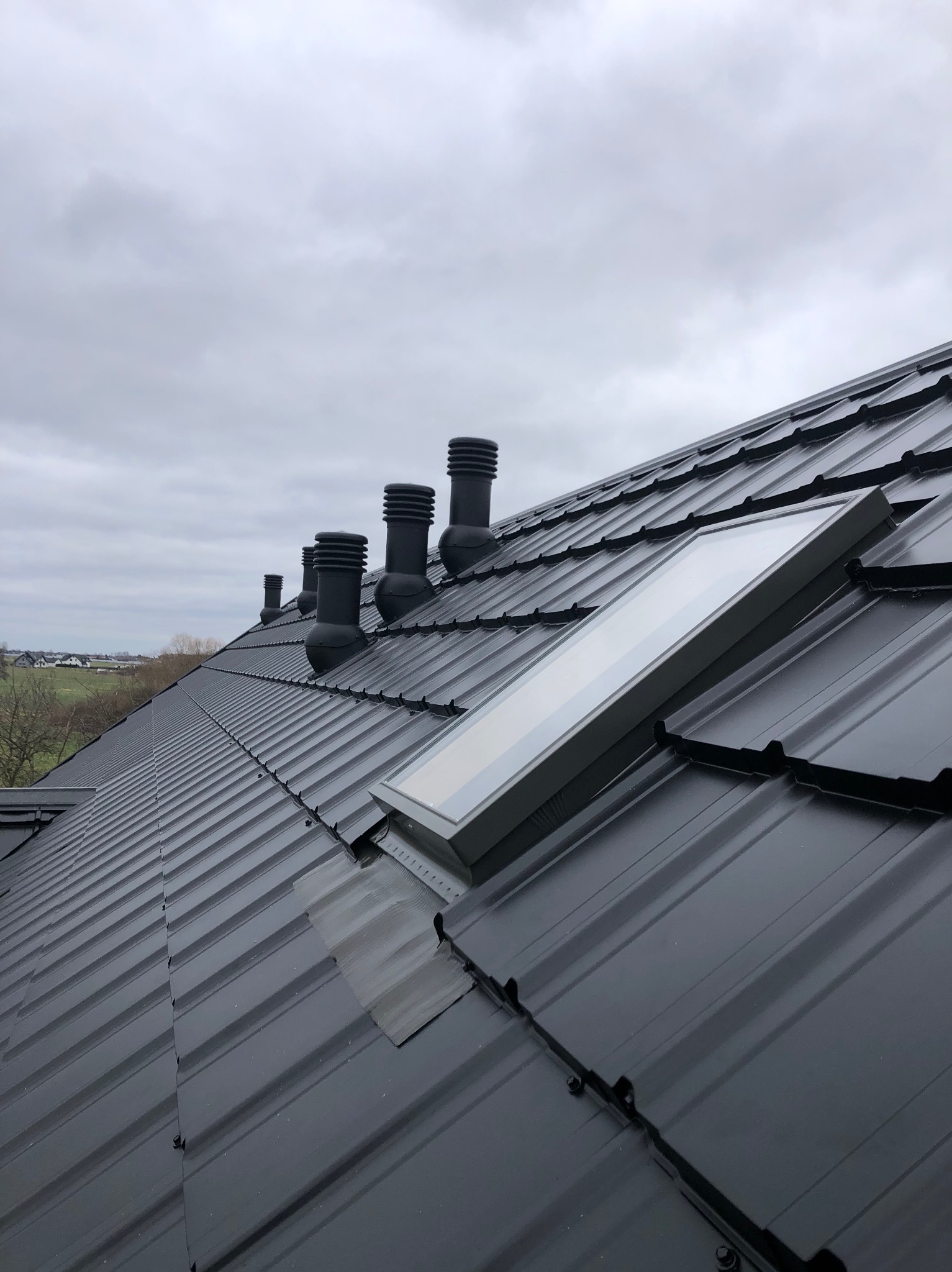 Nowy, czarny dach z blachy trapezowej z oknem dachowym i kominkami wentylacyjnymi na tle pochmurnego nieba.
