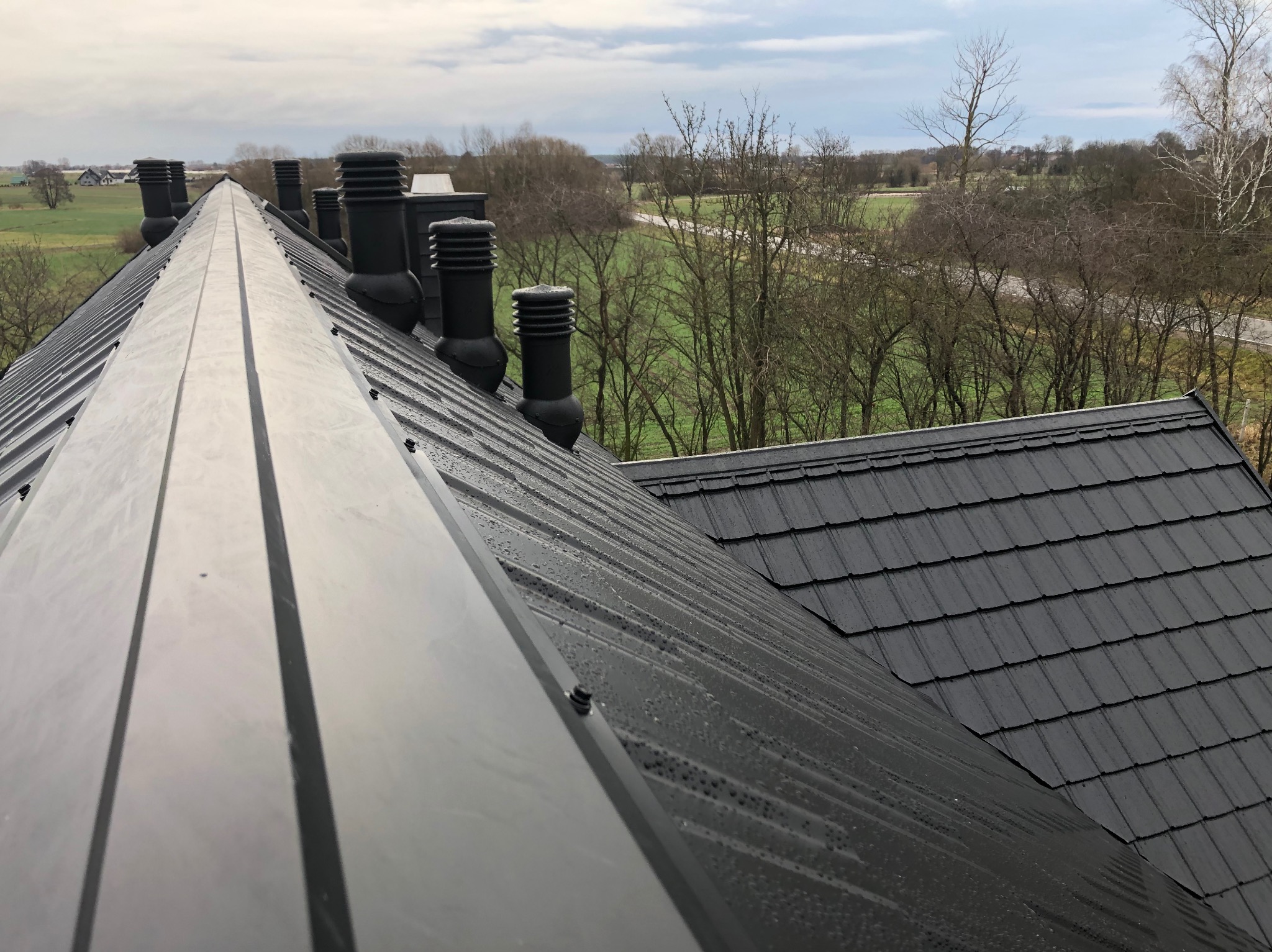 Nowy, czarny dach z blachy na tle pochmurnego nieba i zieleni, widoczne kominy wentylacyjne i krople wody na powierzchni.