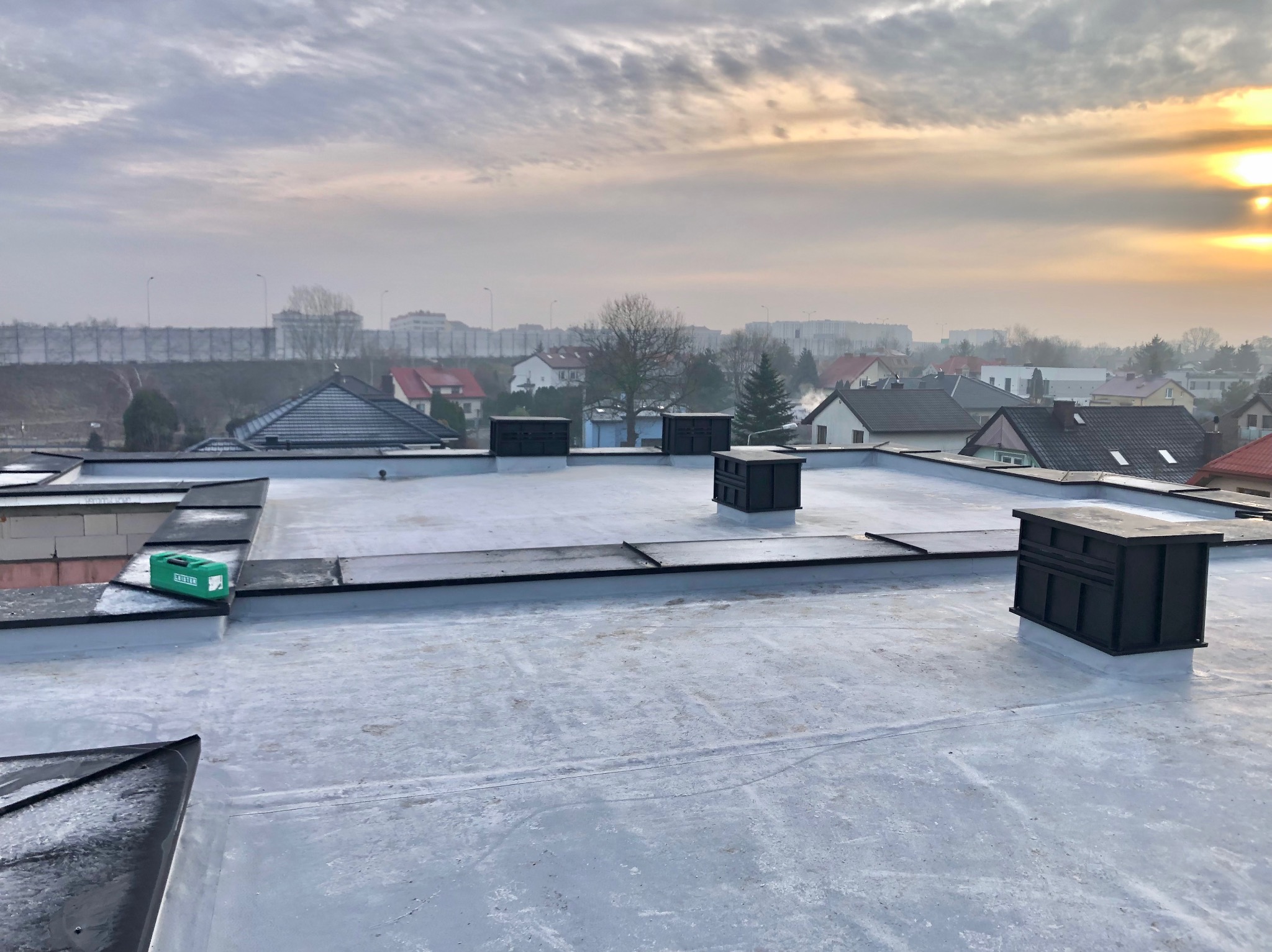 Szary płaski dach z czterema czarnymi kominkami wentylacyjnymi na tle miejskiego krajobrazu o wschodzie słońca. Na dachu widoczny zielony przyrząd pomiarowy.