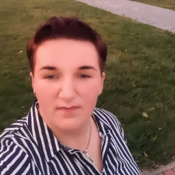 Portret kobiety w pasiastą koszulę, z krótkimi, rudymi włosami i srebrnym naszyjnikiem z krzyżykiem, na tle trawnika i budynków.