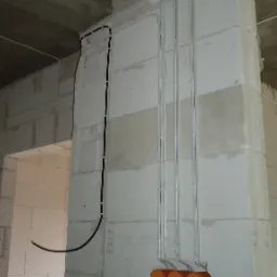 Surowe wnętrze: pionowe rury instalacyjne w ścianie z betonu komórkowego, pomarańczowe puszki elektryczne w dole, czarny kabel biegnący wzdłuż ściany, widoczne oznaczenia wysokości.