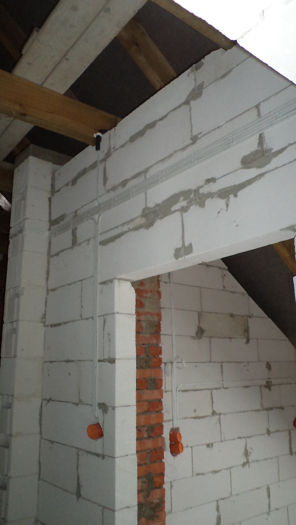 Wewnętrzna ściana z białych bloczków z widocznymi, szarymi przewodami elektrycznymi w peszlach i pomarańczowymi puszkami, fragment więźby dachowej w tle.