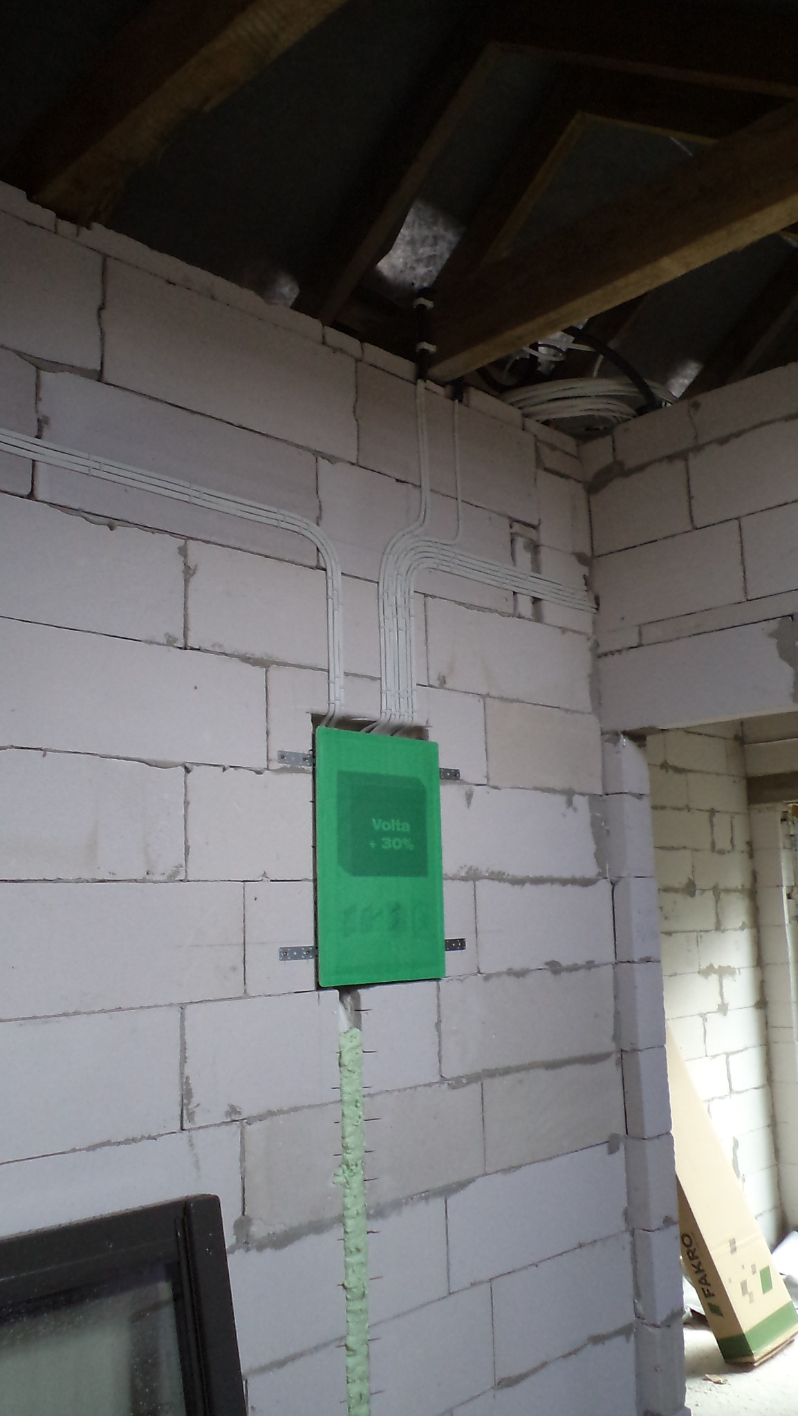 Wewnętrzna instalacja elektryczna w surowym stanie, widoczne przewody prowadzone po ścianie z betonu komórkowego, zabezpieczone zieloną osłoną Volta, oraz ślady pianki montażowej w pionowej...