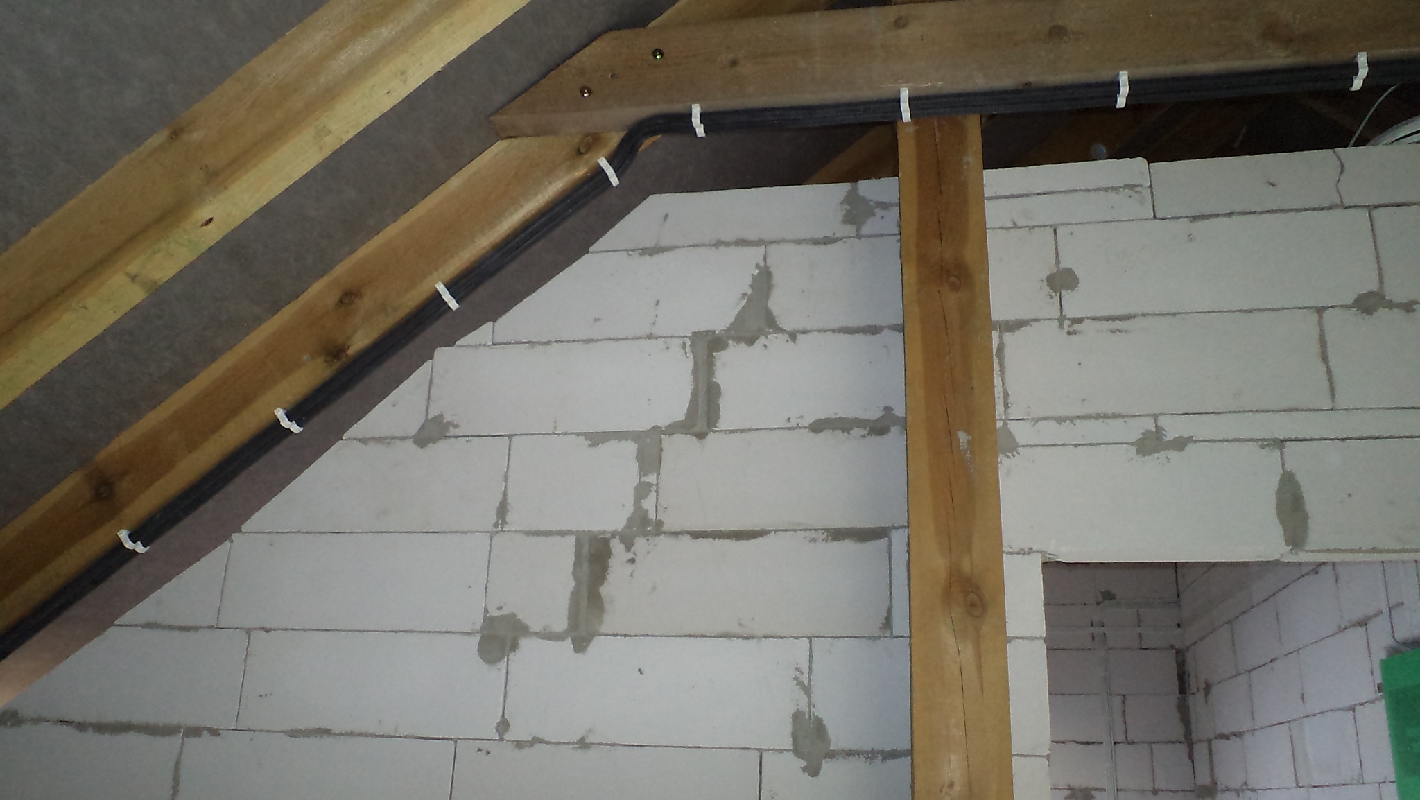 Kabel elektryczny w peszlu przymocowany klamrami do drewnianej konstrukcji poddasza, ściany z betonu komórkowego w tle.