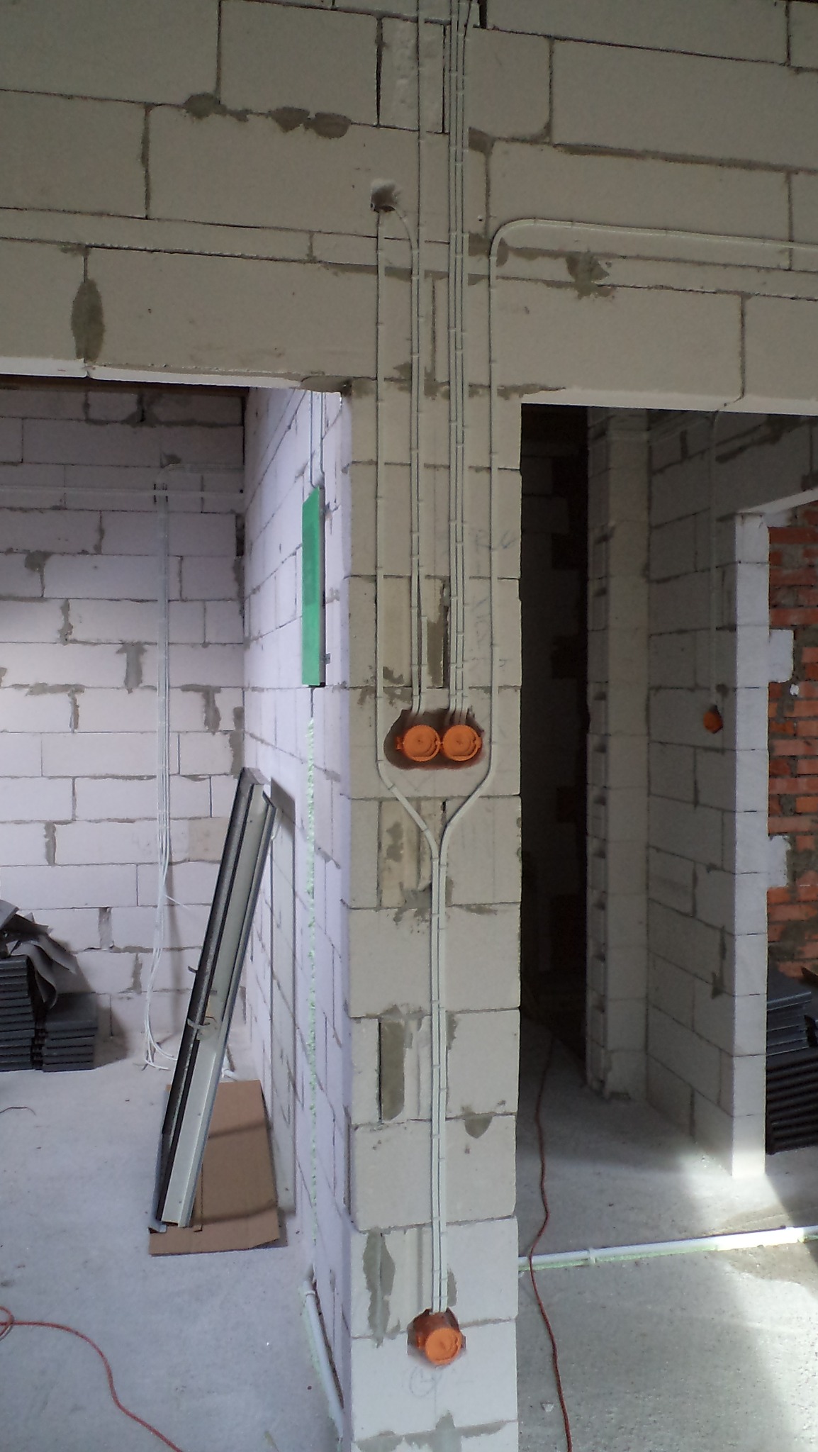 Wewnętrzne okablowanie elektryczne w surowym budynku z bloków betonowych, widoczne pomarańczowe puszki instalacyjne i białe kable prowadzone po ścianach.