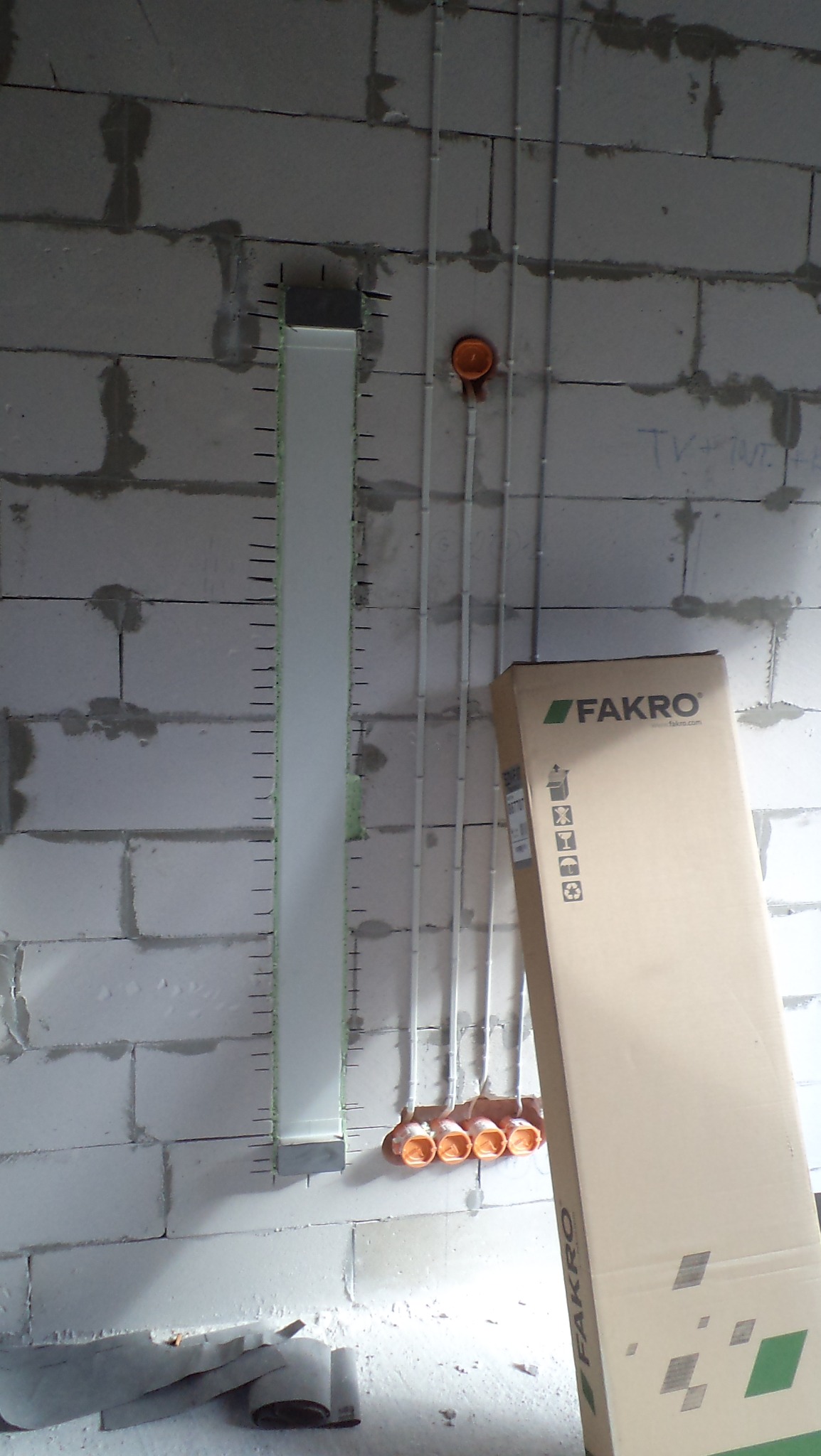 Ściana z betonu komórkowego w trakcie prac instalacyjnych: pionowy kanał instalacyjny, rurki PCV i puszki elektryczne, obok karton Fakro.