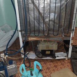 Id-Serwice Sp. z o.o. - Tył białej lodówki z widocznym agregatem, podłączone manometry i butla z czynnikiem chłodniczym na tle wzorzystej podłogi, sugerujące proces serwisowy.