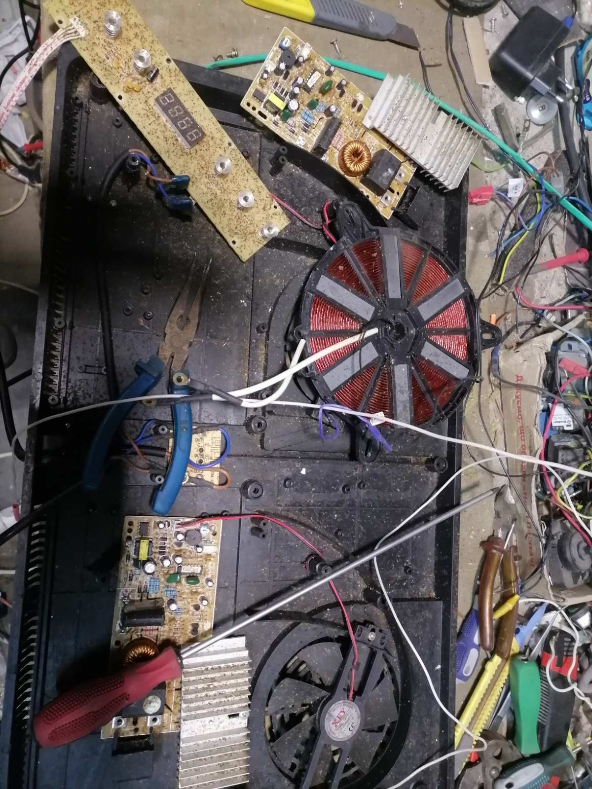 Rozłożona płyta indukcyjna z widocznymi elementami elektronicznymi, cewką grzewczą i wentylatorem, narzędzia leżące obok wskazują na etap naprawy.