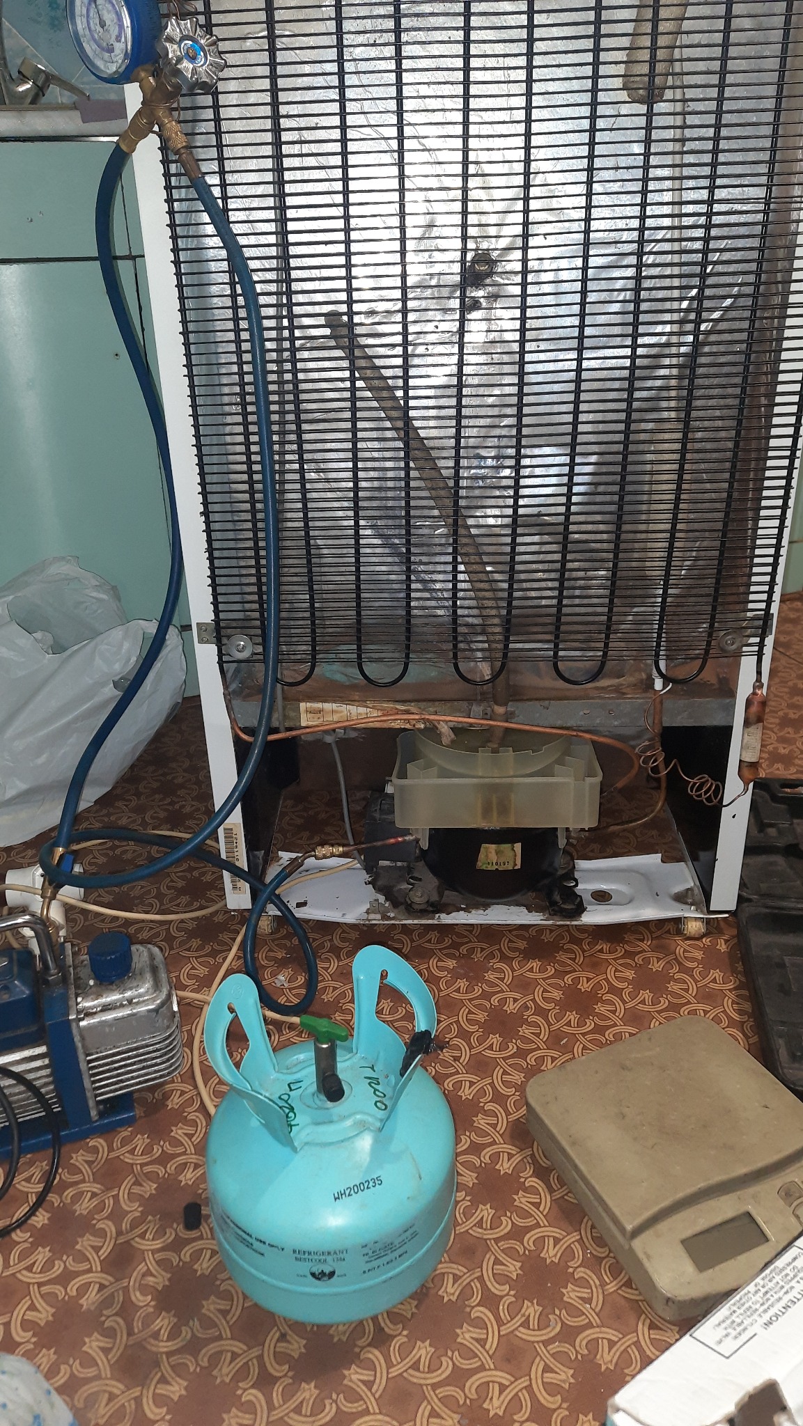 Tył białej lodówki z widocznym agregatem, podłączone manometry i butla z czynnikiem chłodniczym na tle wzorzystej podłogi, sugerujące proces serwisowy.