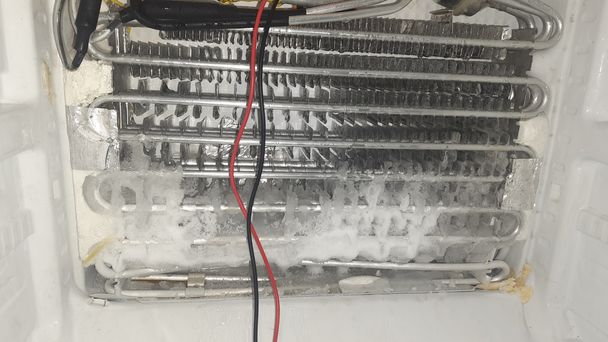 Zamrożony parownik lodówki z widocznymi przewodami elektrycznymi i fragmentami lodu, wnętrze urządzenia z bliska.