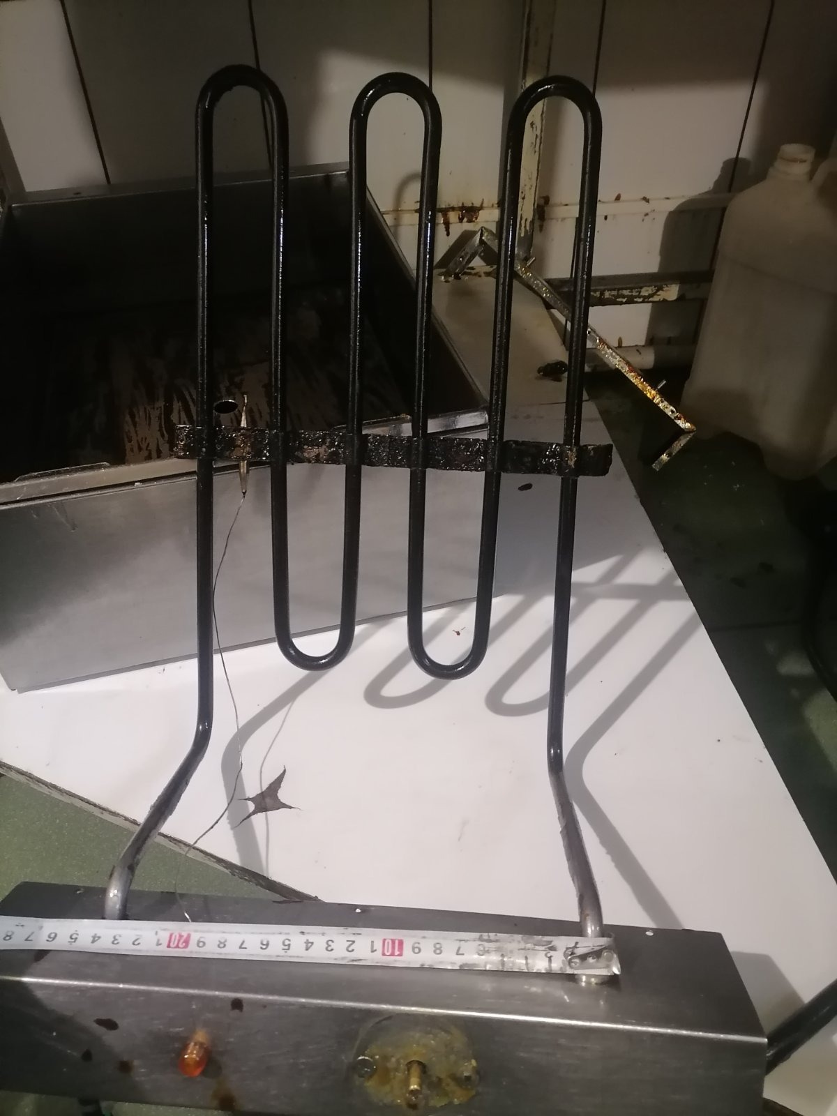 Wyjęta grzałka z piekarnika, widoczne ślady przypaleń i zabrudzeń, obok leży miarka, tło stanowi biały blat i wnętrze piekarnika.