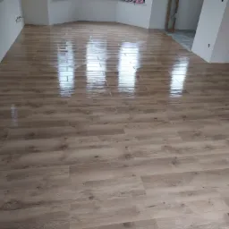 Świeżo położona, błyszcząca podłoga imitująca drewno w jasnym, pustym pomieszczeniu z oknami w wykuszach. Na parapetach resztki materiałów budowlanych.