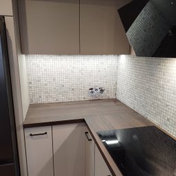 Spartakus PAWEŁ SPALTABAKA - Nowoczesna kuchnia z mozaiką, brązowym blatem i czarną płytą indukcyjną. Widoczne gniazdka elektryczne i oświetlenie LED pod szafkami.