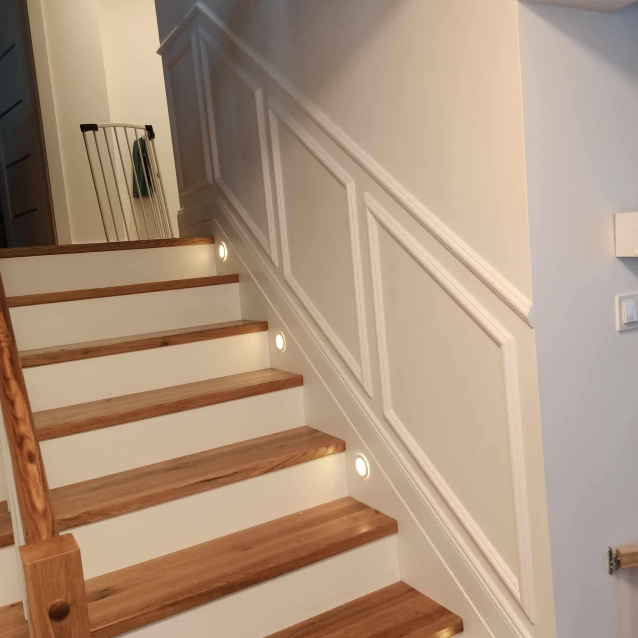 Schody drewniane z białymi podstopnicami i oświetleniem LED w ścianie, dekoracyjne panele ścienne z białymi listwami.