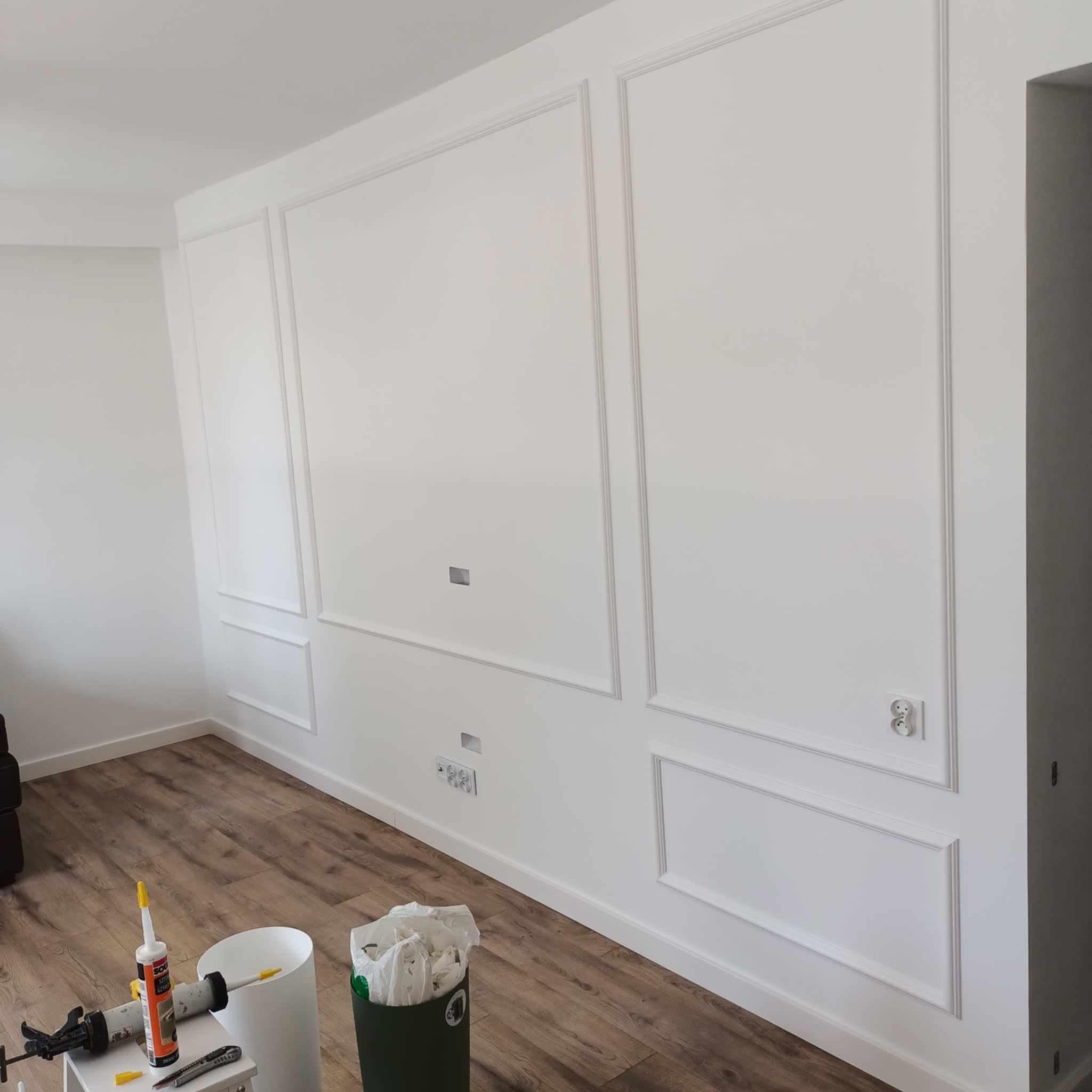 Biała ściana z dekoracyjnymi listwami w stylu klasycznym, widoczne gniazdka elektryczne, nieukończone otwory na kable, narzędzia i resztki materiałów na podłodze.