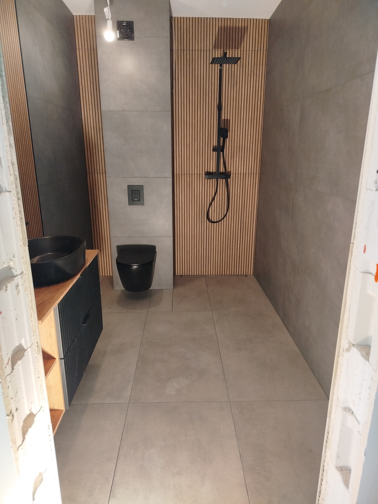 Nowoczesna łazienka z szarymi płytkami, drewnianymi lamelami, czarną armaturą i umywalką nablatową, widok z niedokończonego wejścia.