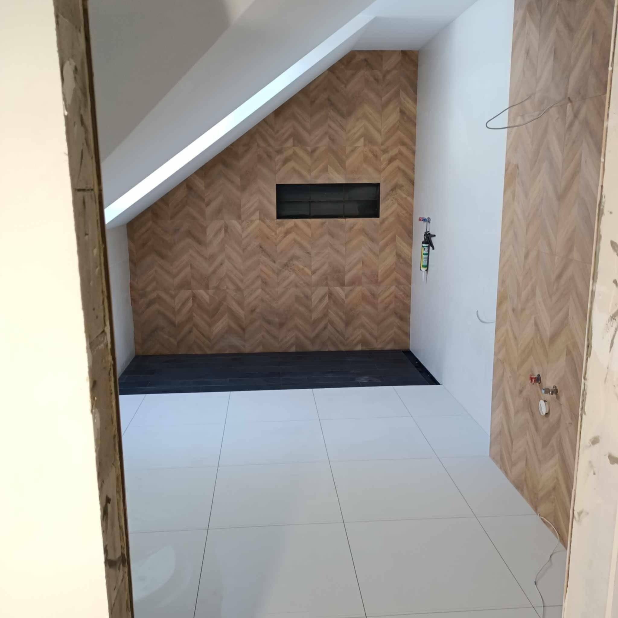 Nowoczesna łazienka na poddaszu z drewnopodobnymi płytkami ułożonymi w jodełkę na ścianie prysznicowej i wnęką z czarnymi płytkami, białe płytki podłogowe, widoczne elementy instalacji wodnej...