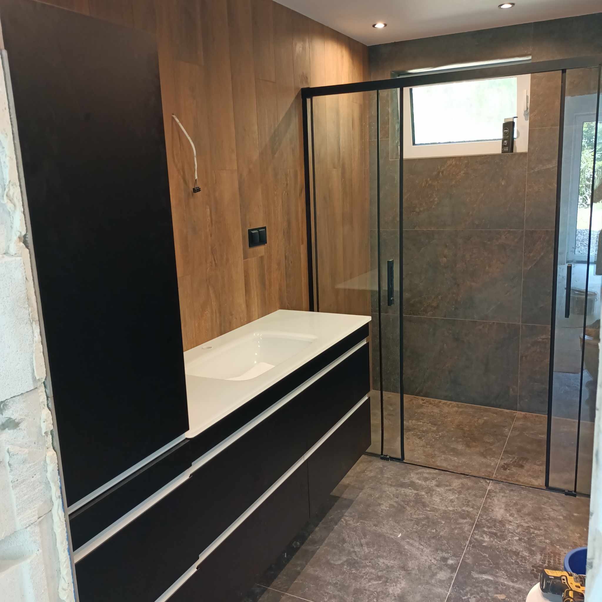 Nowoczesna łazienka w trakcie wykańczania: czarna szafka z umywalką, kabina prysznicowa z czarnymi ramami, ściany wyłożone płytkami imitującymi drewno i kamień, widoczne narzędzia pracy.