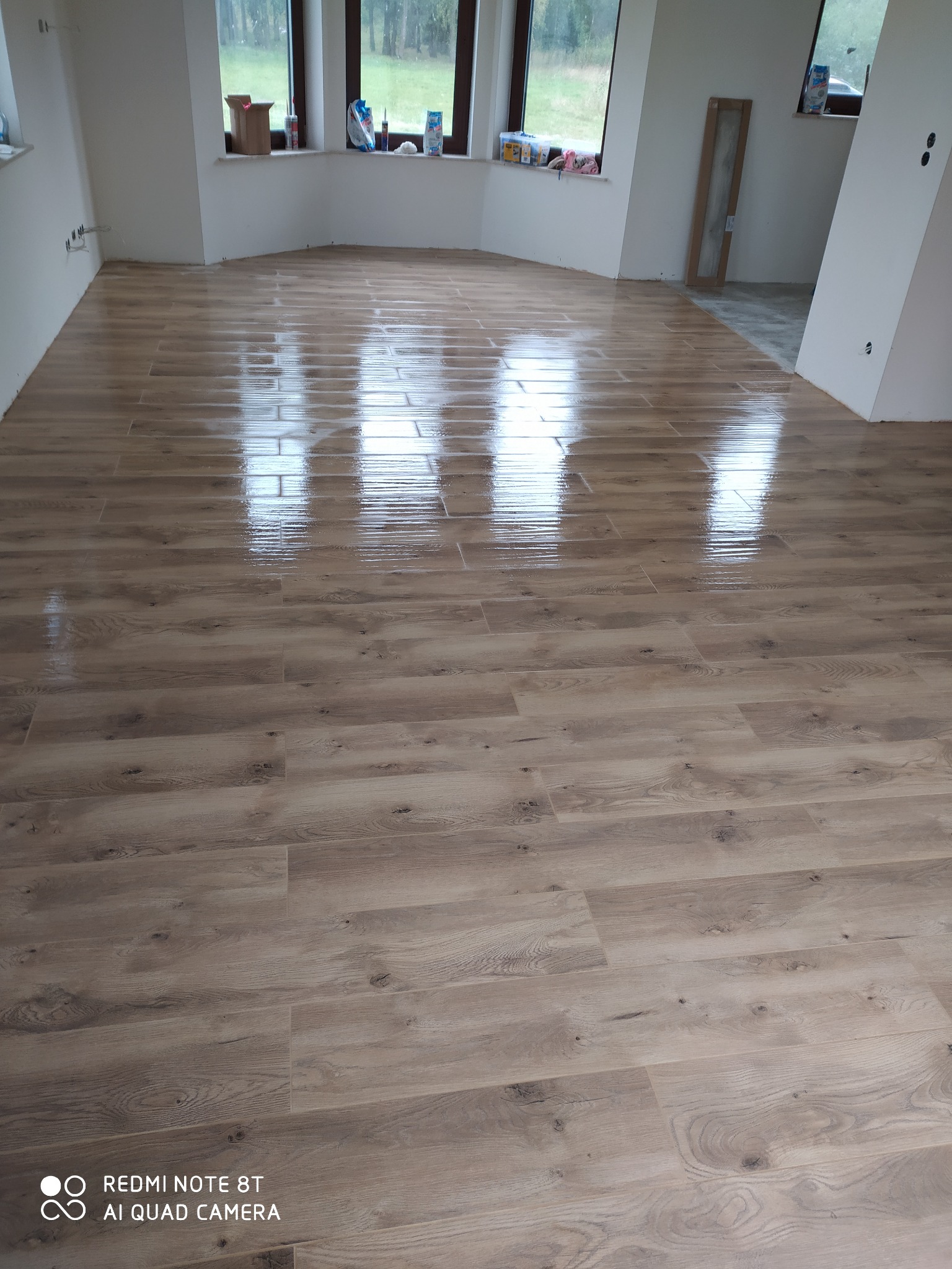 Świeżo położona, błyszcząca podłoga imitująca drewno w jasnym, pustym pomieszczeniu z oknami w wykuszach. Na parapetach resztki materiałów budowlanych.