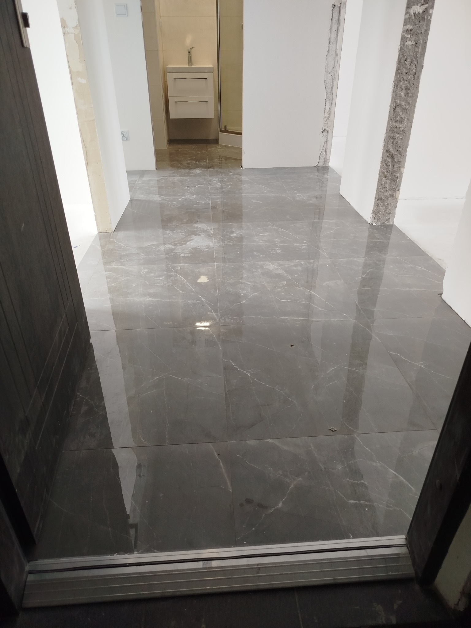 Połyskująca, szara podłoga z płytek imitujących marmur w trakcie remontu łazienki. Widoczne niedokończone ściany i fragment szafki z umywalką w tle.