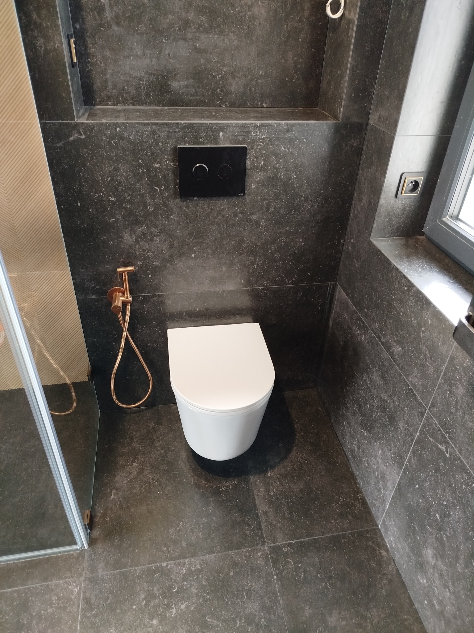 Nowoczesna łazienka z ciemnymi płytkami imitującymi kamień, wiszącą toaletą i złotą armaturą. Minimalistyczny design z akcentem na detale.