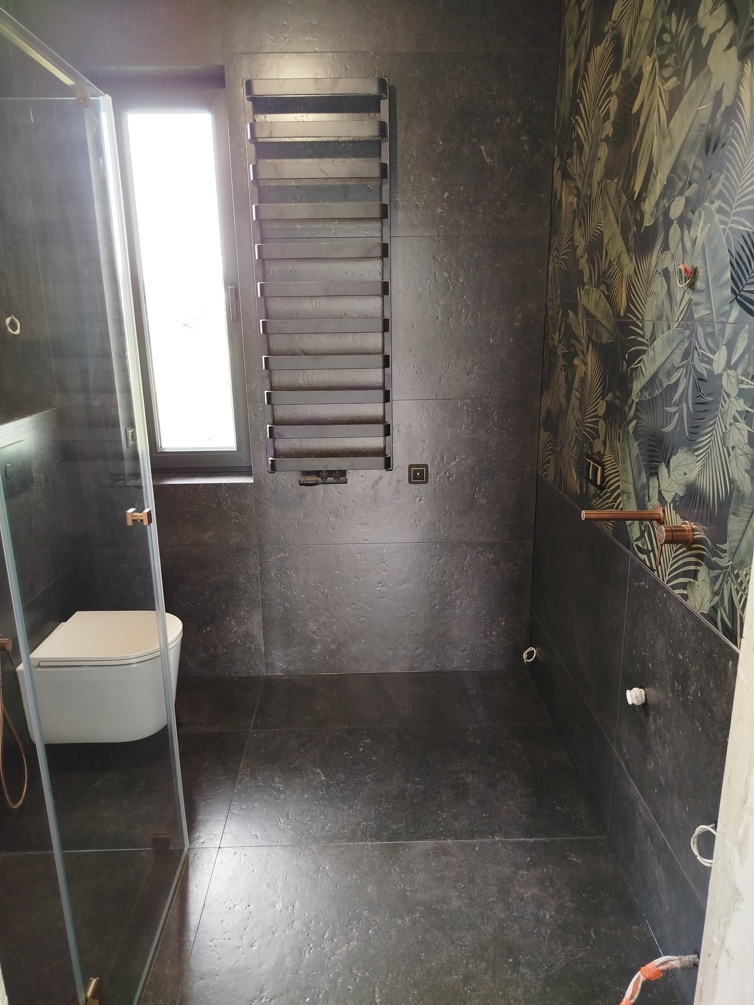 Nowoczesna łazienka z kabiną prysznicową, toaletą, grzejnikiem drabinkowym i ścianą w tropikalne wzory. Ciemna tonacja płytek i złote akcenty armatury.