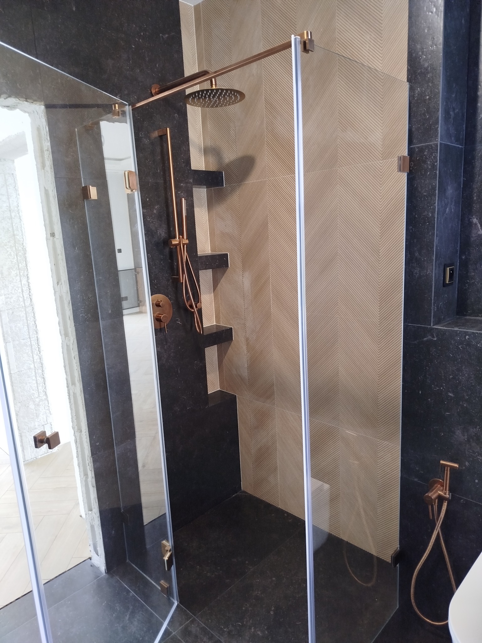 Nowoczesna kabina prysznicowa z deszczownicą w odcieniach beżu i czerni, wykończona elementami w kolorze miedzi. Widoczne geometryczne wzory na płytkach i minimalistyczne półki.