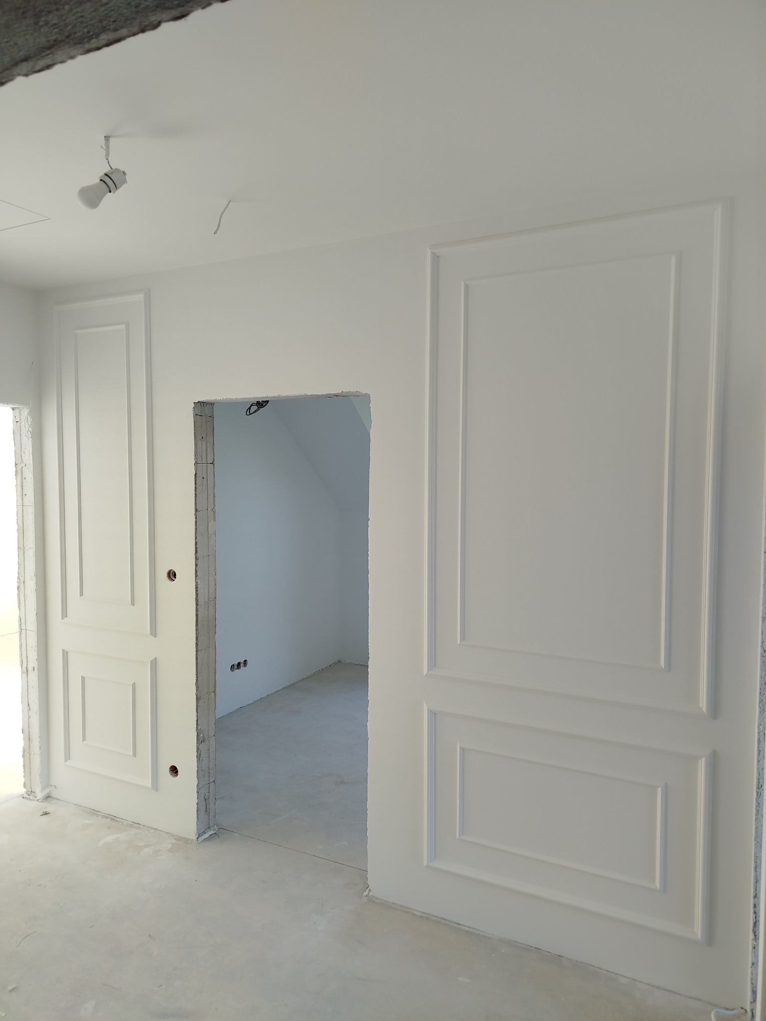 Białe, dekoracyjne panele ścienne z subtelnymi wgłębieniami, tworzące elegancki efekt w nowym wnętrzu. Widoczne surowe wykończenie otworu drzwiowego i niedokończona podłoga.