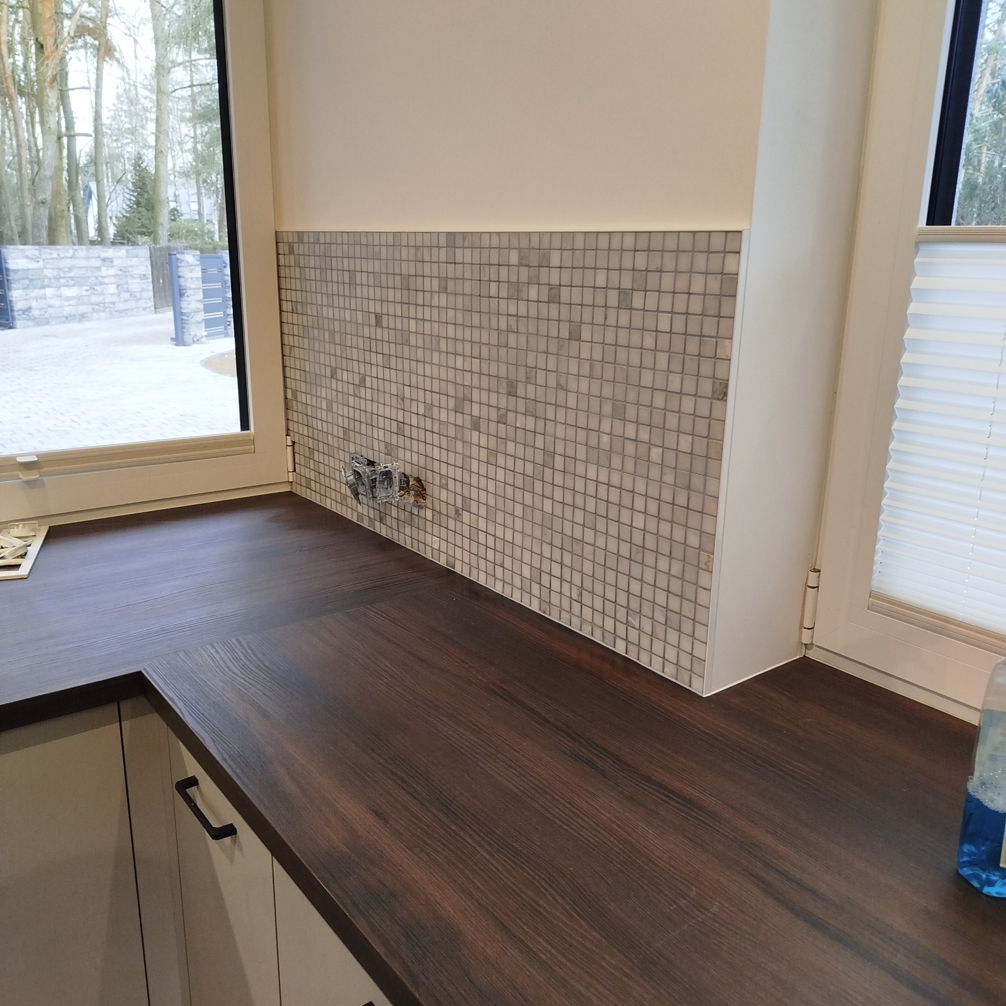 Mozaika z drobnych płytek na ścianie nad blatem, widoczne gniazdka elektryczne przed wykończeniem. Ujęcie z perspektywy blatu, z oknem w tle.