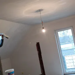 Poddasze w trakcie prac wykończeniowych: szpachlowanie sufitu przez pracownika, widoczne rusztowanie, wiadro z masą szpachlową Knauf Rotband Finish oraz surowa żarówka na kablu.