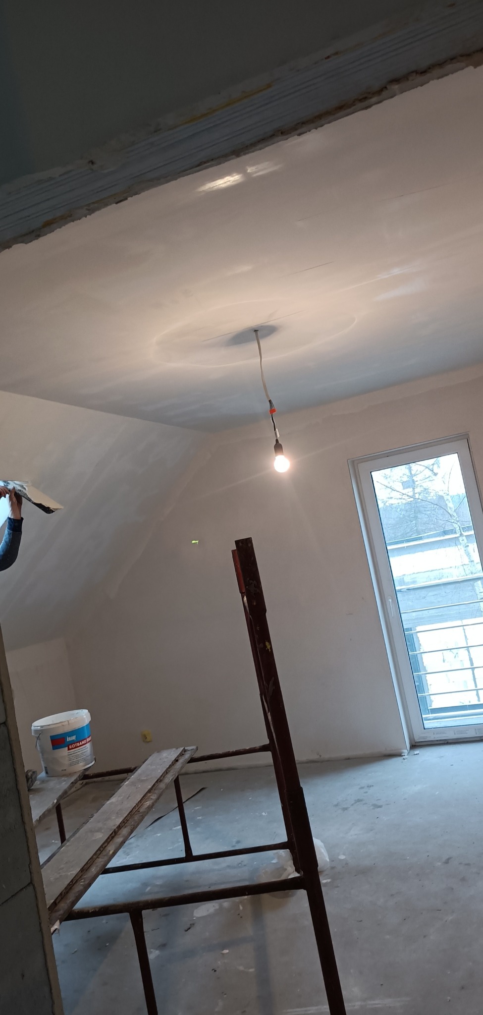 Poddasze w trakcie prac wykończeniowych: szpachlowanie sufitu przez pracownika, widoczne rusztowanie, wiadro z masą szpachlową Knauf Rotband Finish oraz surowa żarówka na kablu.