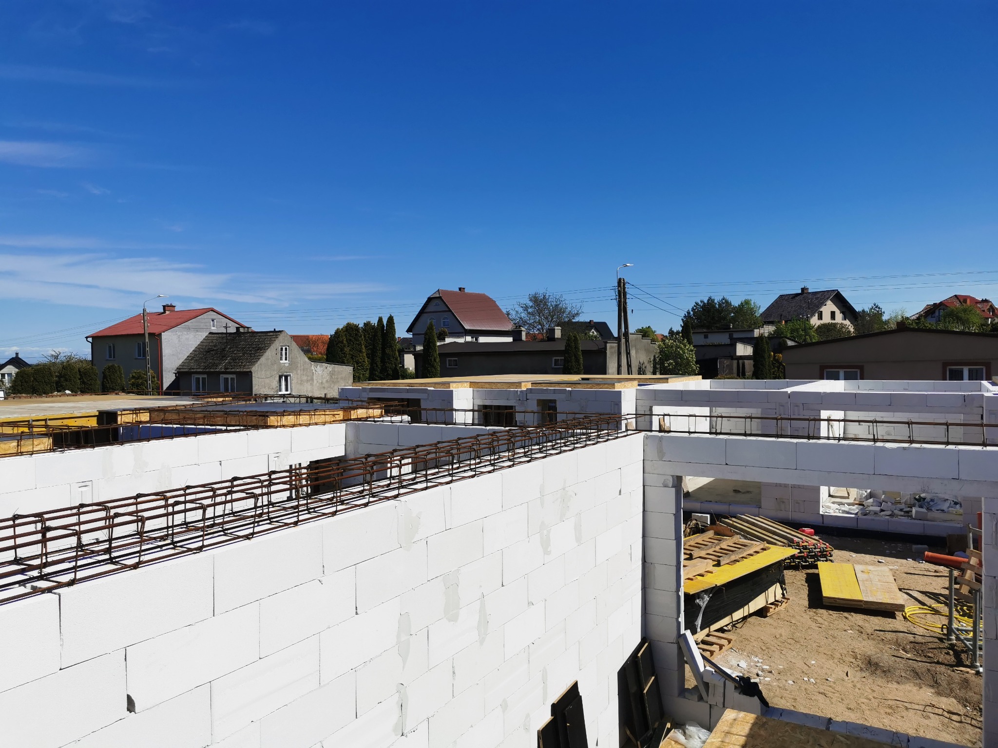 Budowa domu jednorodzinnego w stanie surowym otwartym: białe bloczki, zbrojenie, szalunki, widok na okoliczne domy i błękitne niebo.