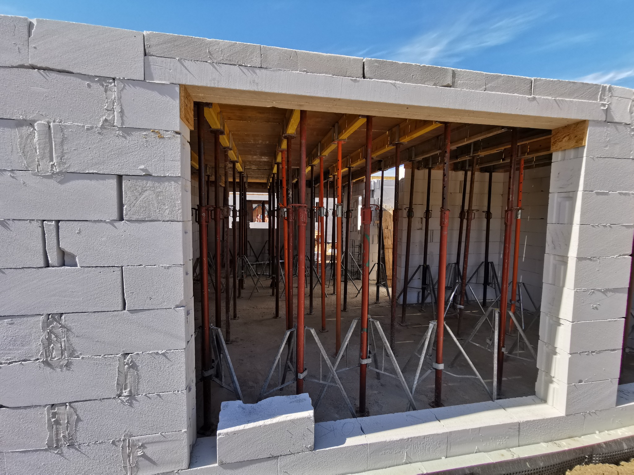 Wznoszenie ściany z białych bloczków betonu komórkowego Ytong, widoczne stemple budowlane podtrzymujące strop, niebo w tle.