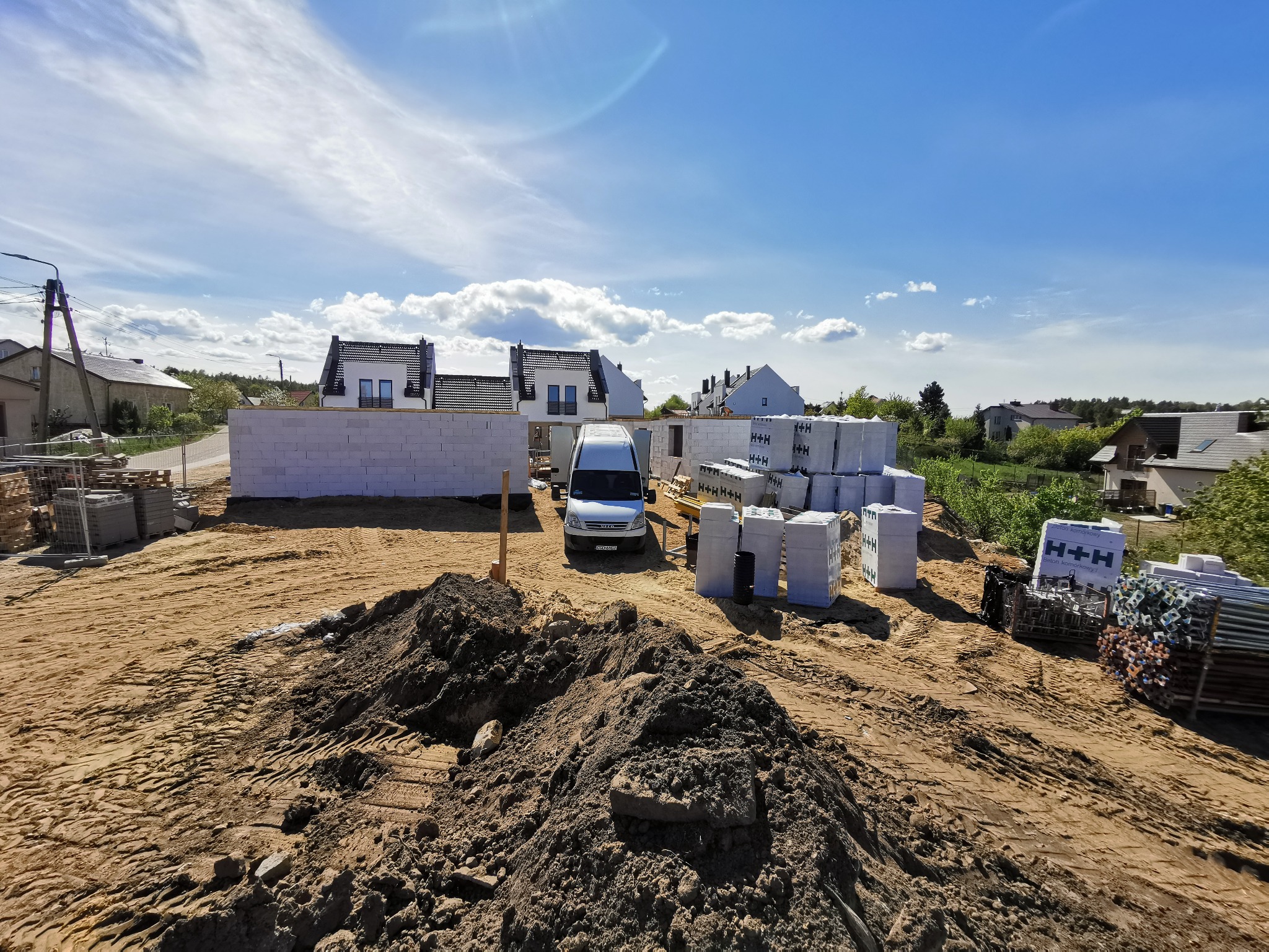Budowa domów jednorodzinnych w trakcie wznoszenia ścian z bloczków betonowych, z widocznymi paletami materiałów budowlanych i samochodem dostawczym na placu budowy, w tle zabudowa mieszkalna...