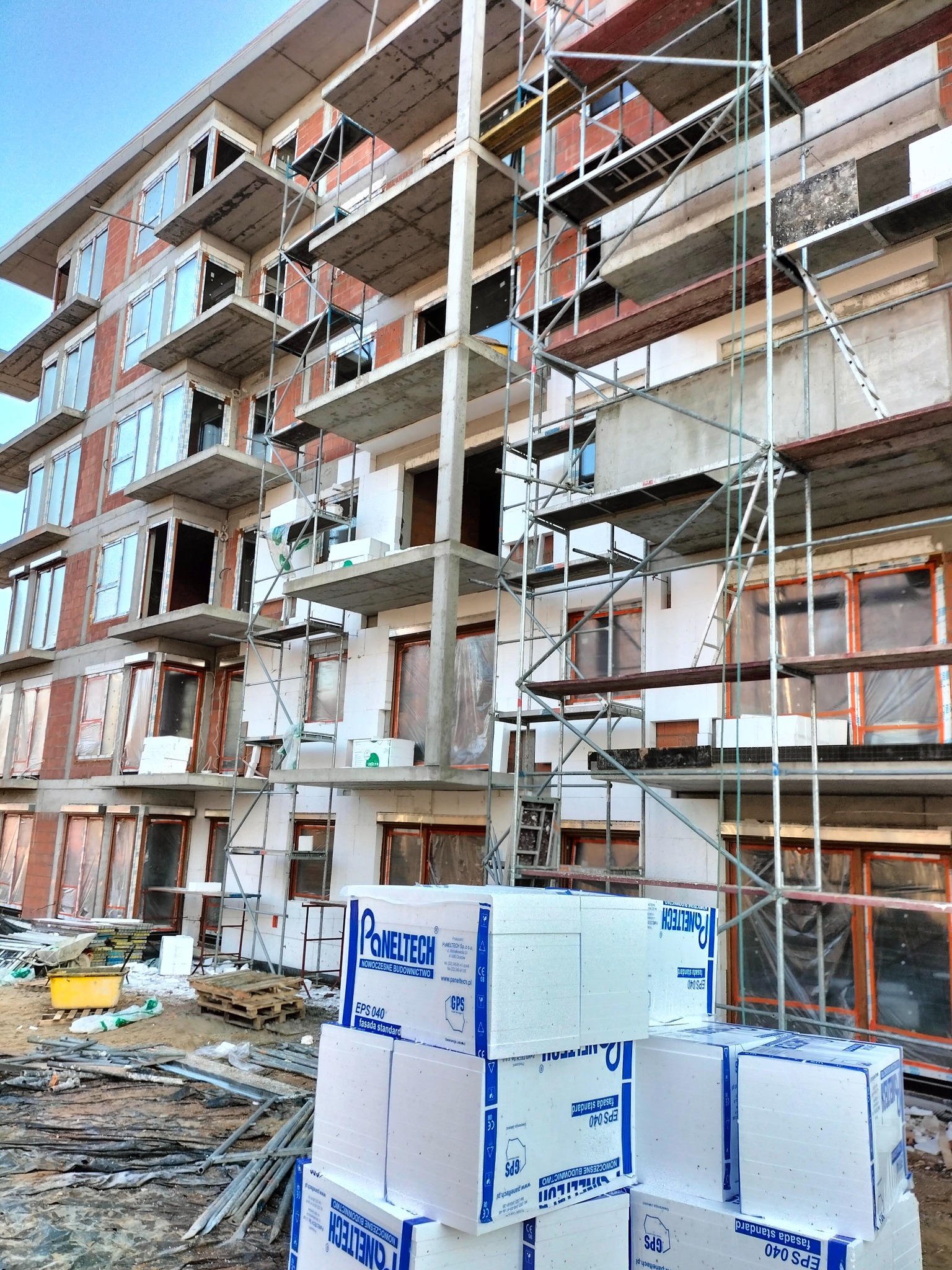 Wielopiętrowy budynek w trakcie ocieplania, widoczne rusztowania, niezabudowane okna, oraz stos paczek styropianu EPS 040 z logo Paneltech na pierwszym planie.