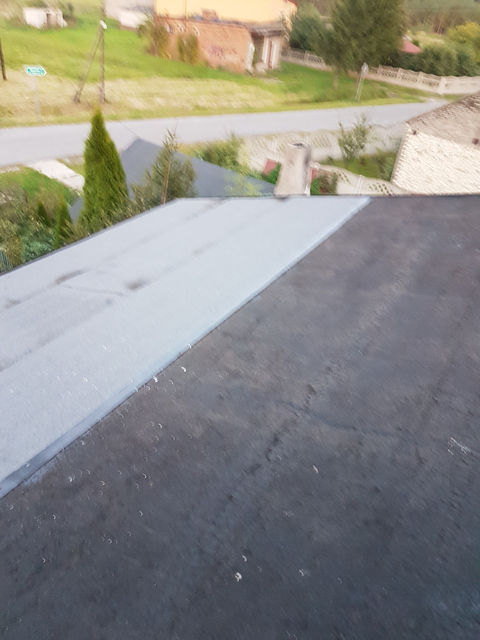 Widok z góry na fragment dachu po hydroizolacji w Gidlach, z widocznym połączeniem starej i nowej powłoki, w tle zabudowania mieszkalne i zieleń.