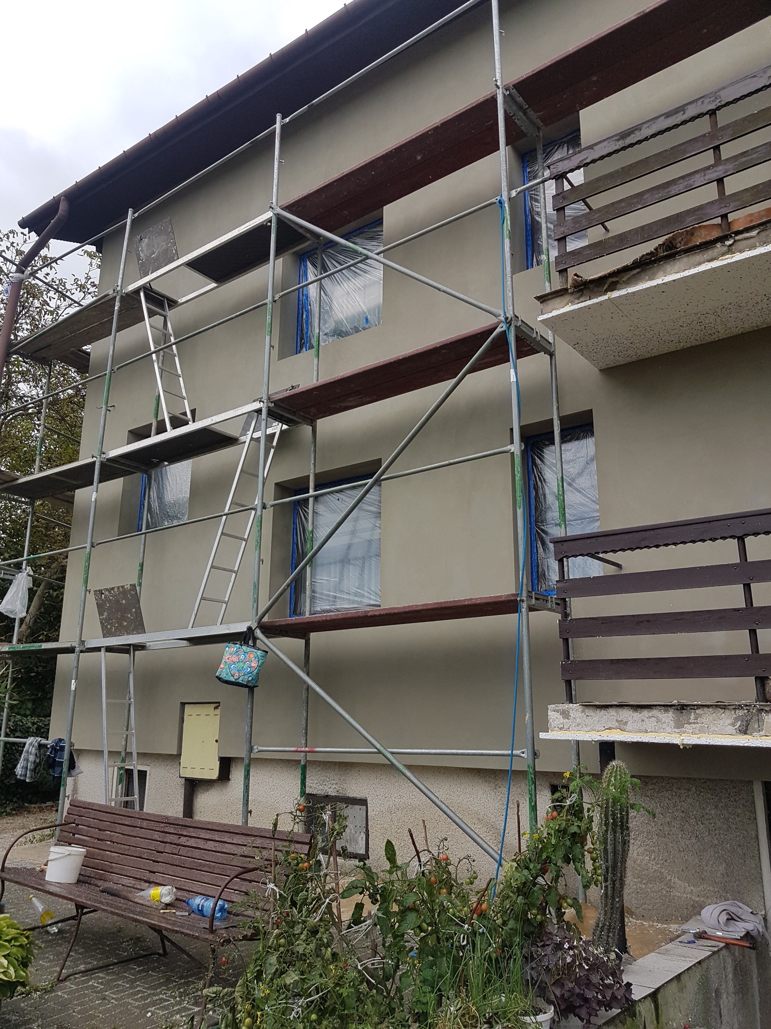 Dom jednorodzinny w trakcie tynkowania elewacji, okna zabezpieczone folią, widoczne rusztowanie, na pierwszym planie ławka z wiaderkiem i torba zawieszona na rusztowaniu.