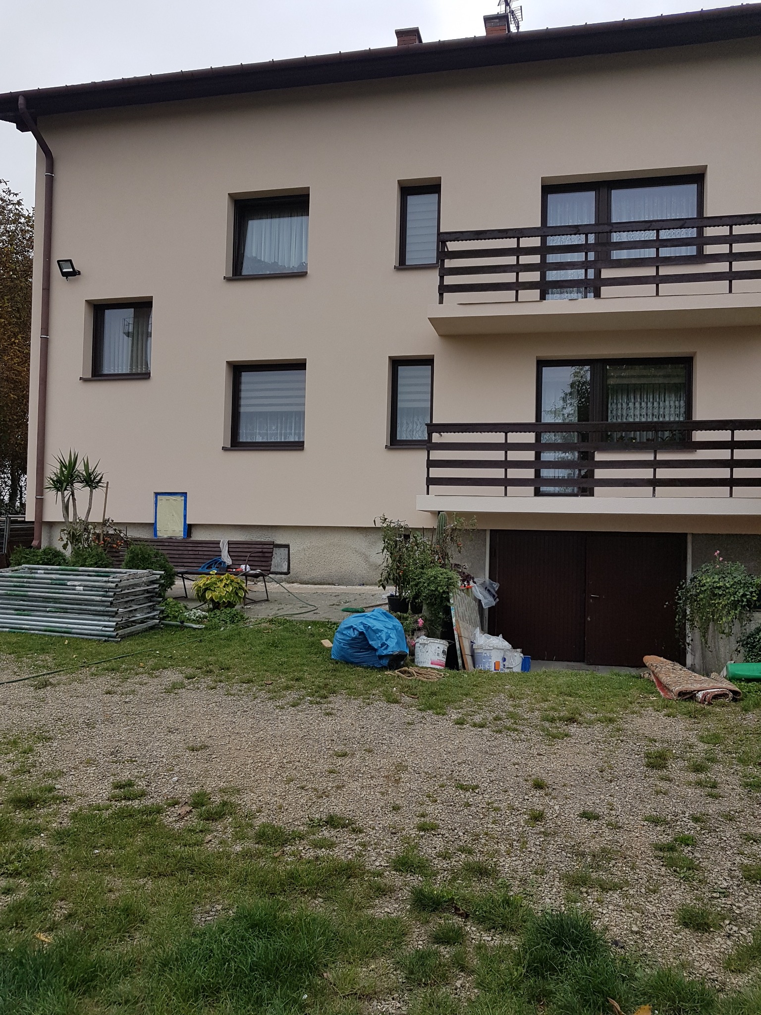 Dwupiętrowy dom z beżową elewacją, ciemnymi oknami i balkonem, z widocznymi rusztowaniami i materiałami budowlanymi na trawniku przed budynkiem, sugerującymi prace wykończeniowe.