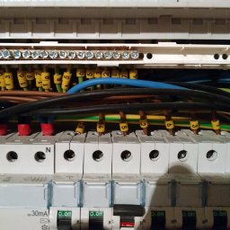 Sebastian Wioska - Szczegółowe ujęcie wnętrza skrzynki elektrycznej z widocznymi bezpiecznikami, przewodami oznaczonymi numerami w żółtych koszulkach i listwą zaciskową ze śrubami.