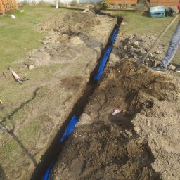 Sebastian Wioska - Wykopany rów w trawniku z niebieską folią zabezpieczającą, widoczne narzędzia i osoba z łopatą, przygotowanie do instalacji podziemnej.