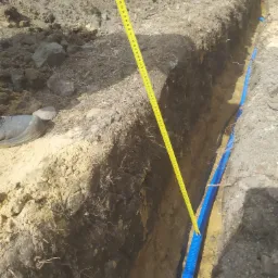 Wykop w ziemi z ułożoną niebieską rurą karbowaną, obok leży miarka zwinięta do pomiaru głębokości.