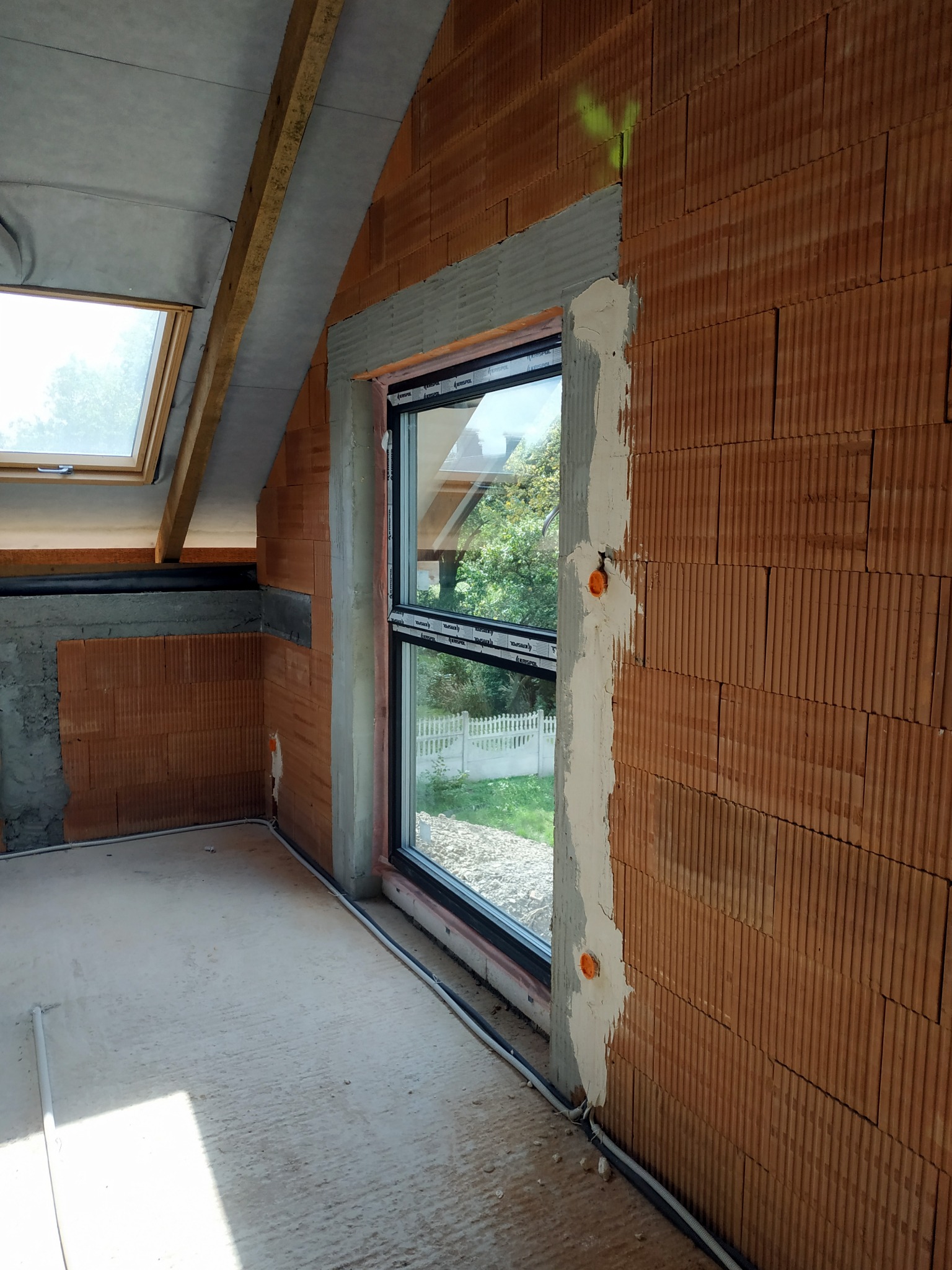 Wnętrze w stanie surowym z oknami dachowymi i pionowymi, ściany z czerwonej cegły z widocznymi przewodami elektrycznymi, częściowo otynkowane otwory okienne.