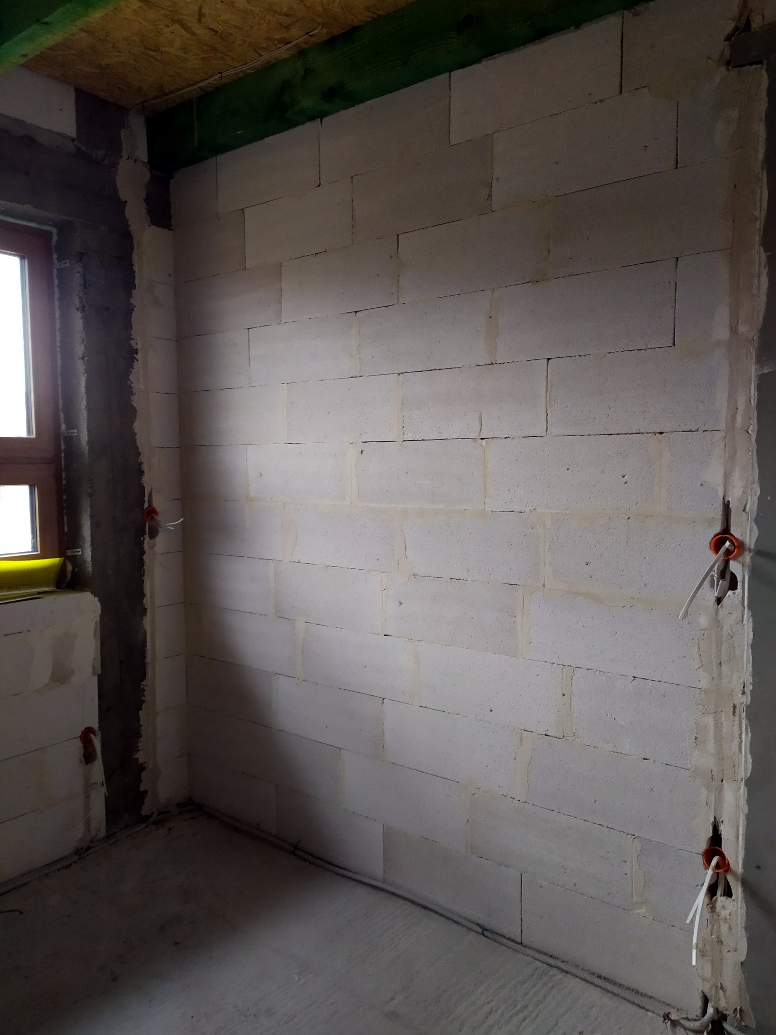 Wnętrze w trakcie budowy: ściana z białych bloczków z widocznymi puszkami instalacyjnymi i przewodami elektrycznymi, surowy beton na podłodze, okno z widocznym żółtym elementem na parapecie.