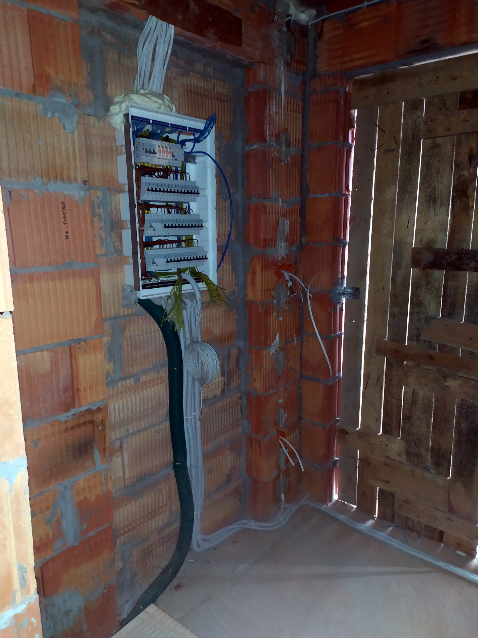 Rozdzielnia elektryczna zamontowana na ceglanej ścianie w surowym stanie, widoczne kable w peszlu i luźne przewody, tymczasowe drzwi z desek.