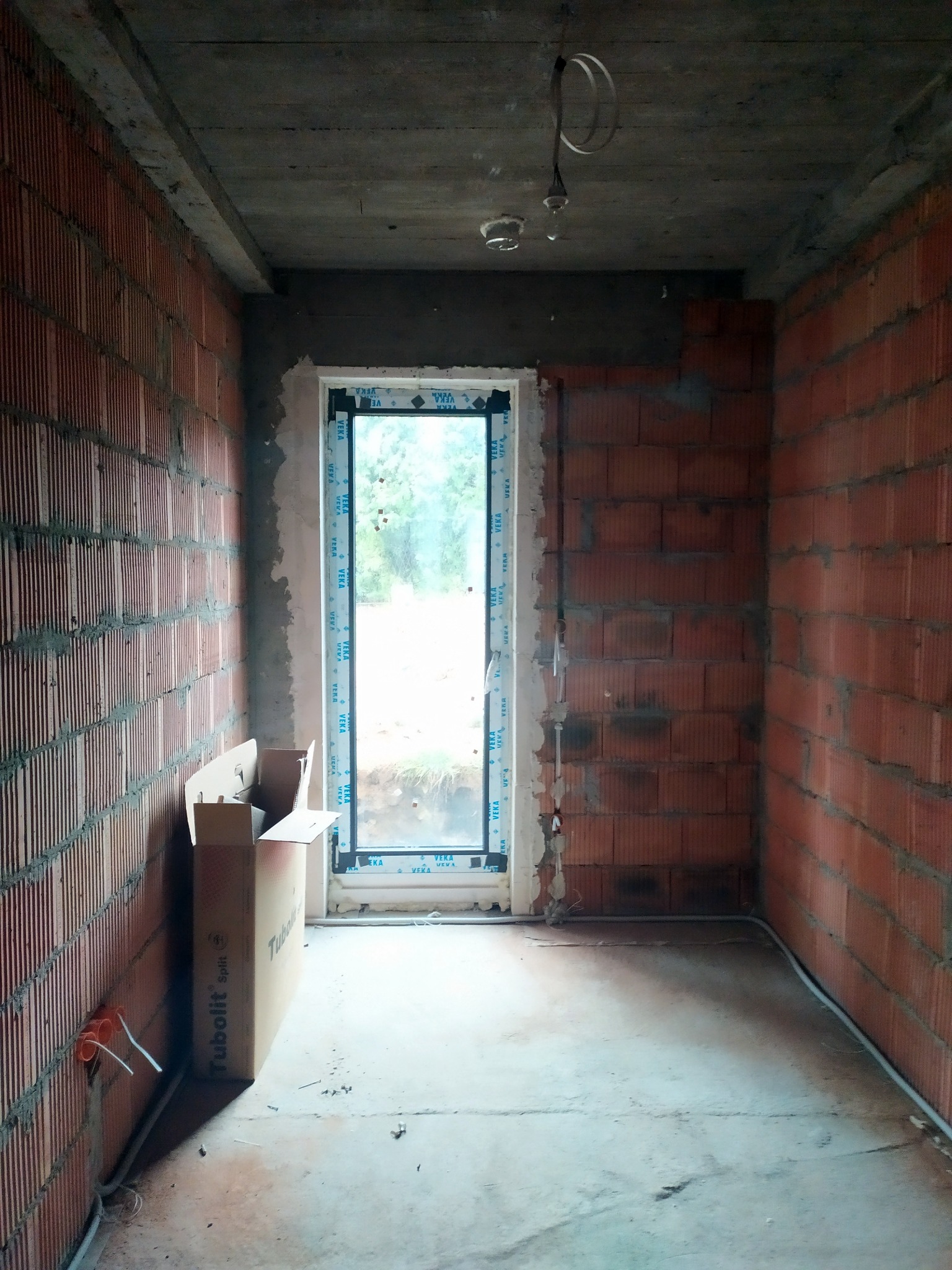 Surowe wnętrze w trakcie budowy: ściany z czerwonej cegły, okno z naklejkami VEKA, wystające przewody elektryczne i karton Tubolit Split.