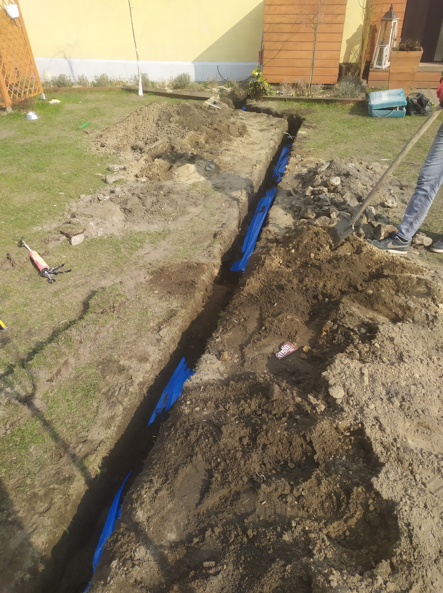 Wykopany rów w trawniku z niebieską folią zabezpieczającą, widoczne narzędzia i osoba z łopatą, przygotowanie do instalacji podziemnej.