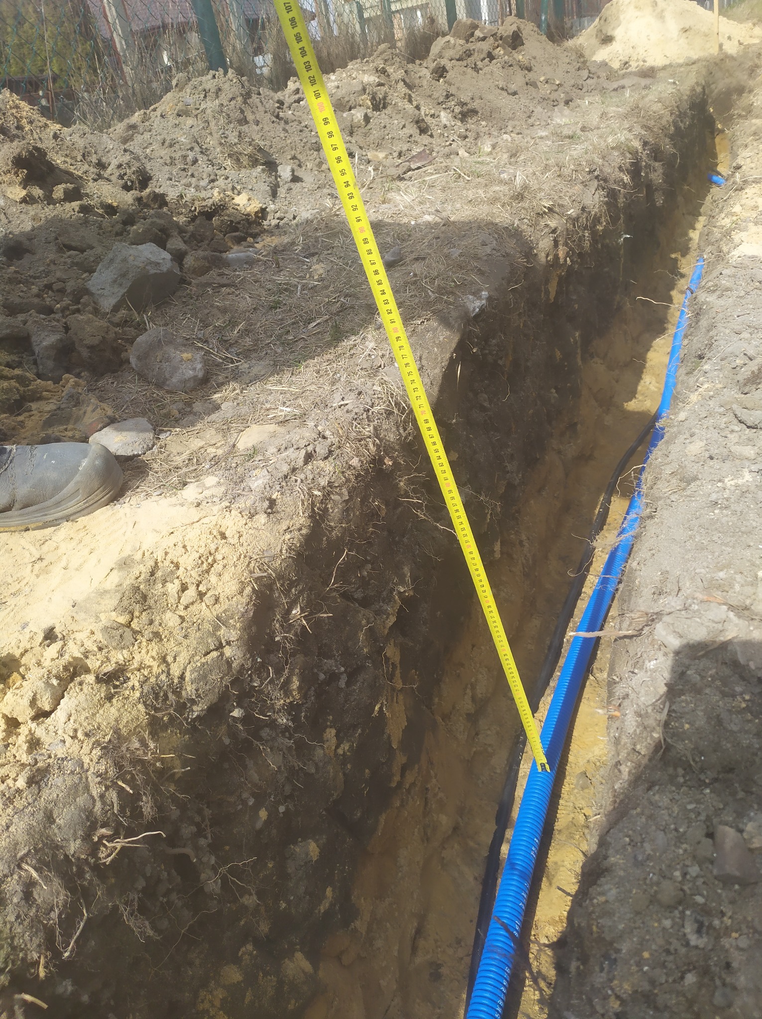 Wykop w ziemi z ułożoną niebieską rurą karbowaną, obok leży miarka zwinięta do pomiaru głębokości.
