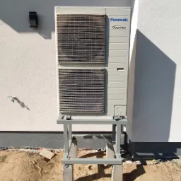 Zewnętrzna jednostka pompy ciepła Panasonic Inverter zamontowana na metalowym stelażu z betonowymi podstawami, przy białej elewacji budynku z widocznym punktem poboru wody i okrągłym otworem...